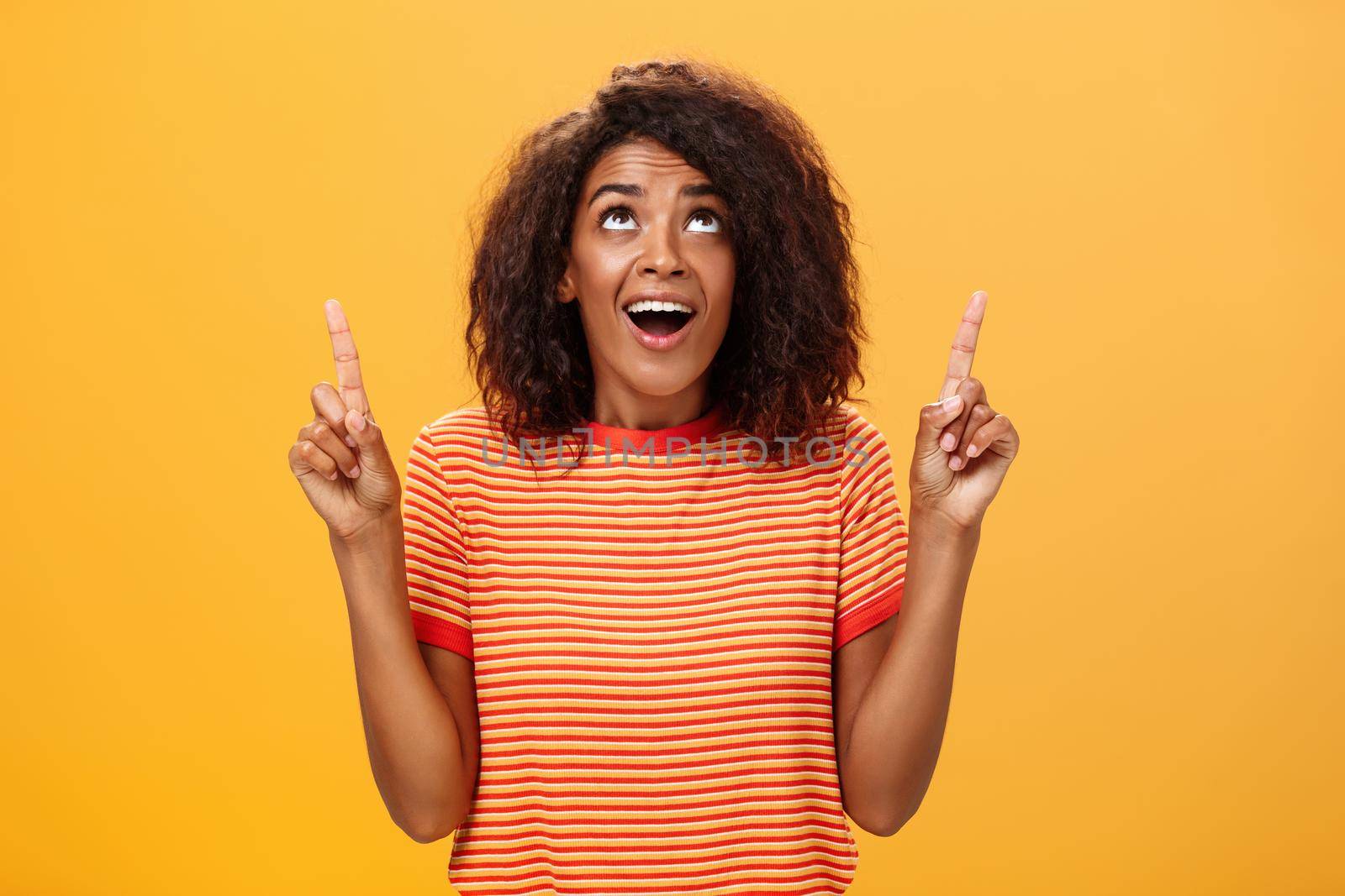 Optimistic happy african american girl saying thanks to god looking and pointing up pleased and carefree starting day in productive way being delighted with lucky chance over orange background. Body language and emotions concept