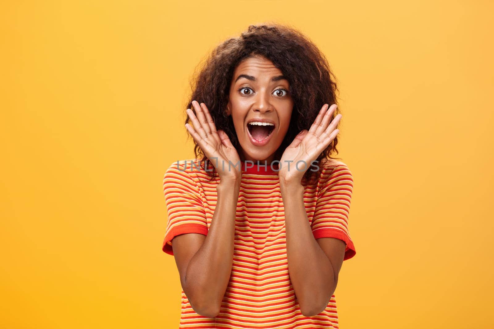 Waist-up shot of impressed excited talkative dark-skinned woman with curly hair and perfect skin yelling from happiness and amazement shaking palms near face cheering for awesome sale over orange wall. Lifestyle.