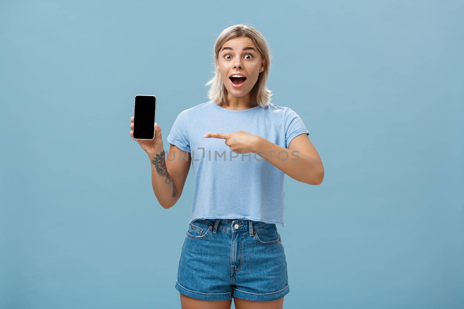 Studio shot of amazed and impressed charming european blonde in casual clothes gasping charmed at camera while showing smartphone screen pointing at device with finger over blue background by Benzoix