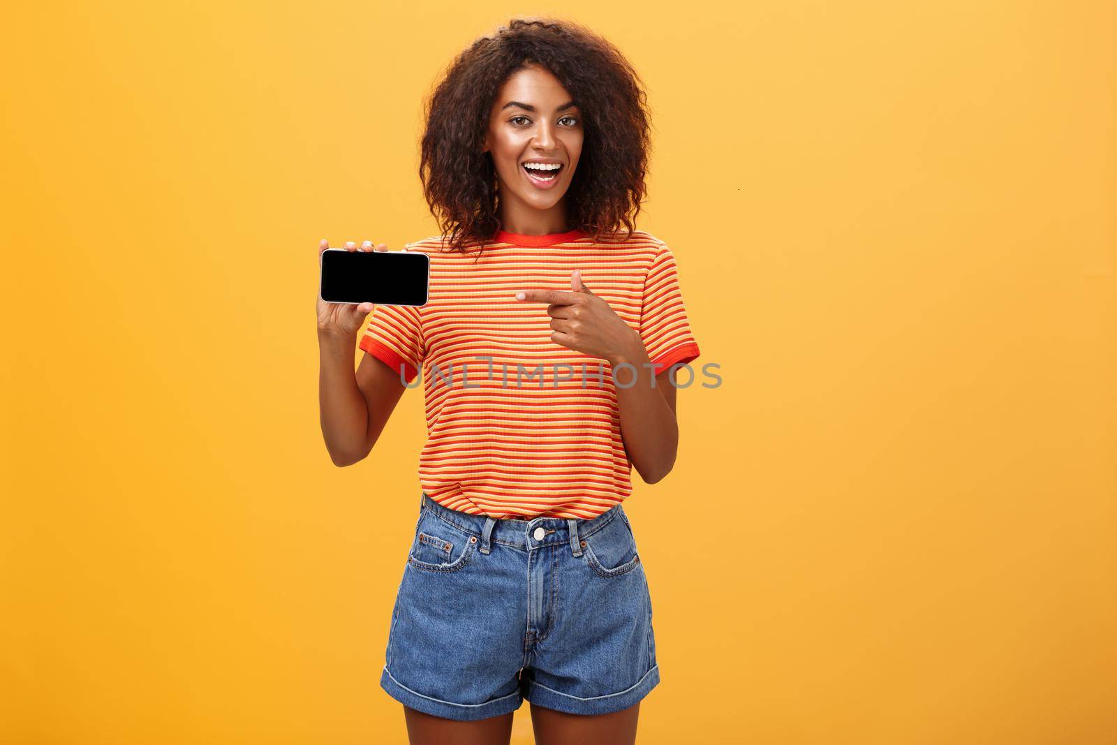 Girl showing picture in phone bragging about her cool vacation. Charming friendly and sociable african american woman with curly hair holding cellphone pointing at smartphone screen over orange wall. Technology and advertisement concept