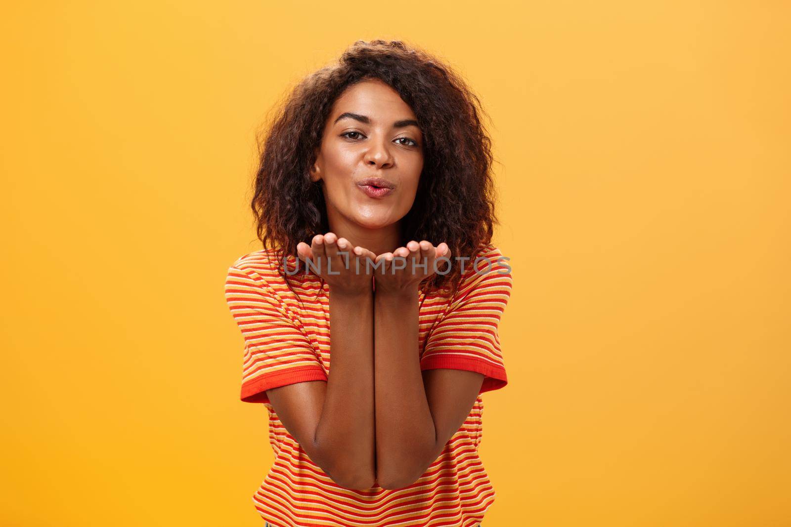 Sending passionate kiss to most loving person. Romantic attractive and stylish young african american girlfriend with curly hairstyle bending towards camera with slight smile, folded lips blowing mwah.