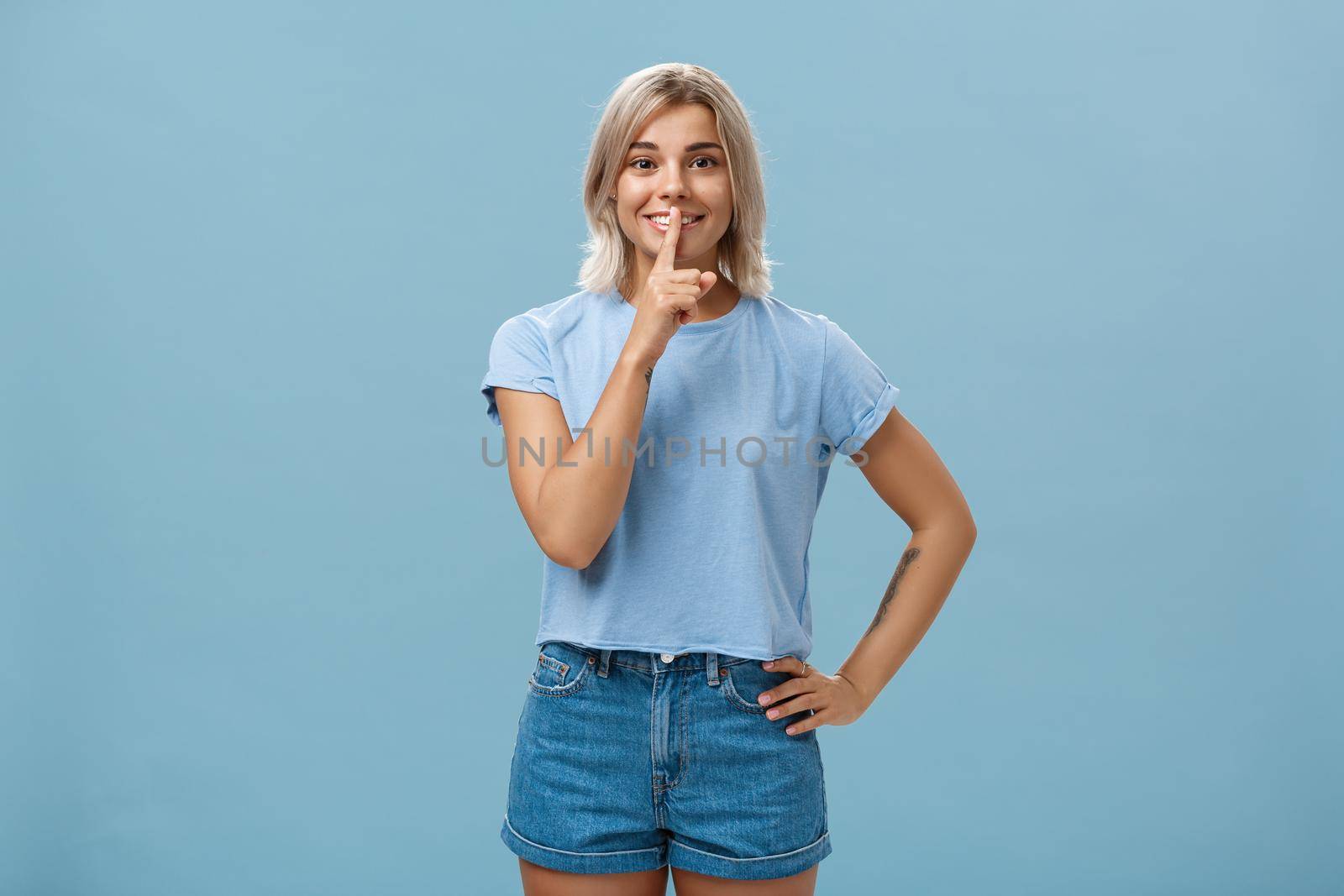 Studio shot of dreamy optimistic good-looking fair-haired female in denim shorts saying shh showing shush gesture with index finger over mouth smiling hiding secret over blue background. Body language concept