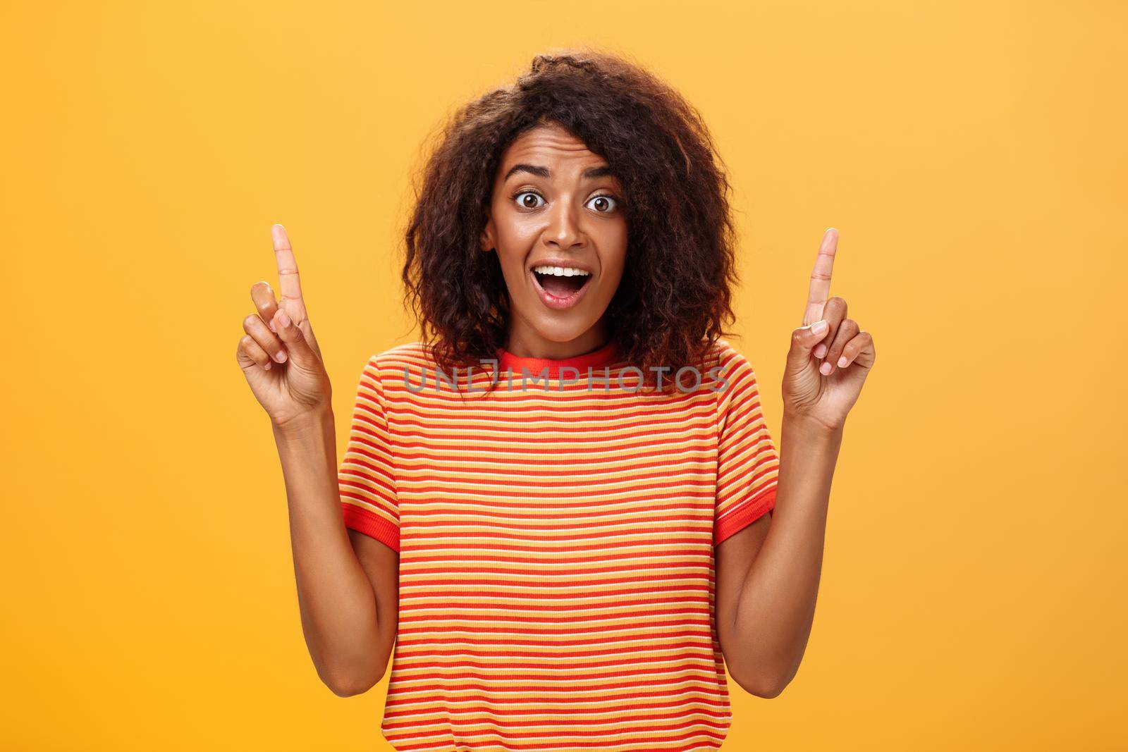 Waist-up shot of amazed happy enthusiastic cute african american woman with curly hairstyle in striped t-shirt raising index fingers pointing up gazing mesmerized and delighted at camera over orange wall. Lifestyle.