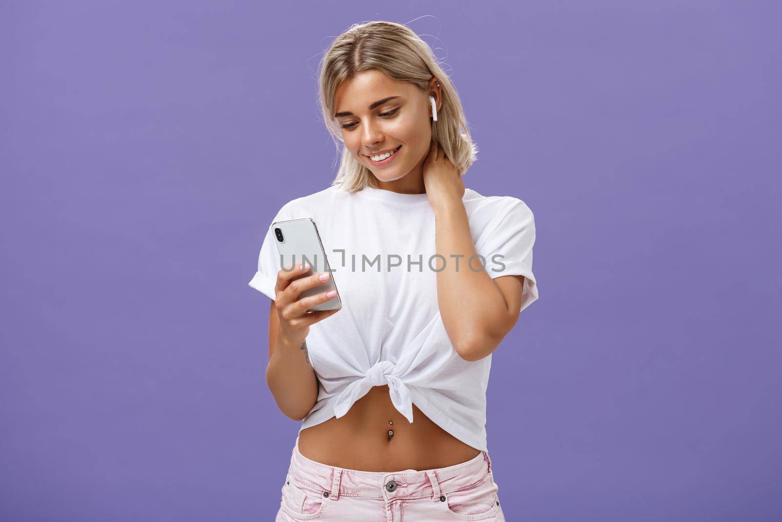 Woman receiving warm romantic message being delighted and touched feeling love gazing at smartphone screen wearing wireless earphones touching neck gently being sensitive and feminine by Benzoix