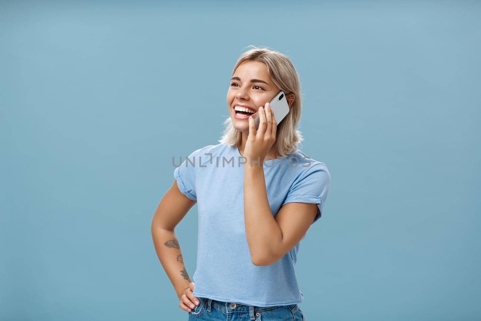 Lifestyle. Waist-up shot of sociable amused and happy attractive caucasian fair-haired woman in casual t-shirt standing half-turned gazing left with hand on hip while talking on smartphone over blue background.