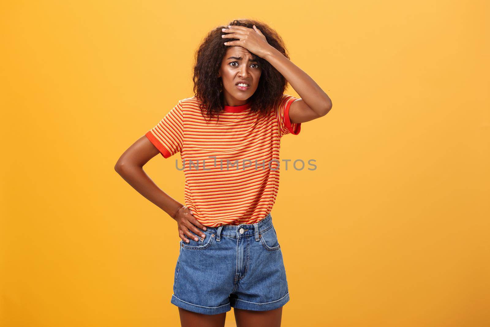 Shit I am in trouble. Concerned anxious cute dark-skinned woman in trendy t-shirt and shorts clenching teeth with guilty look frowning punching forehead having trouble or problem over orange wall.