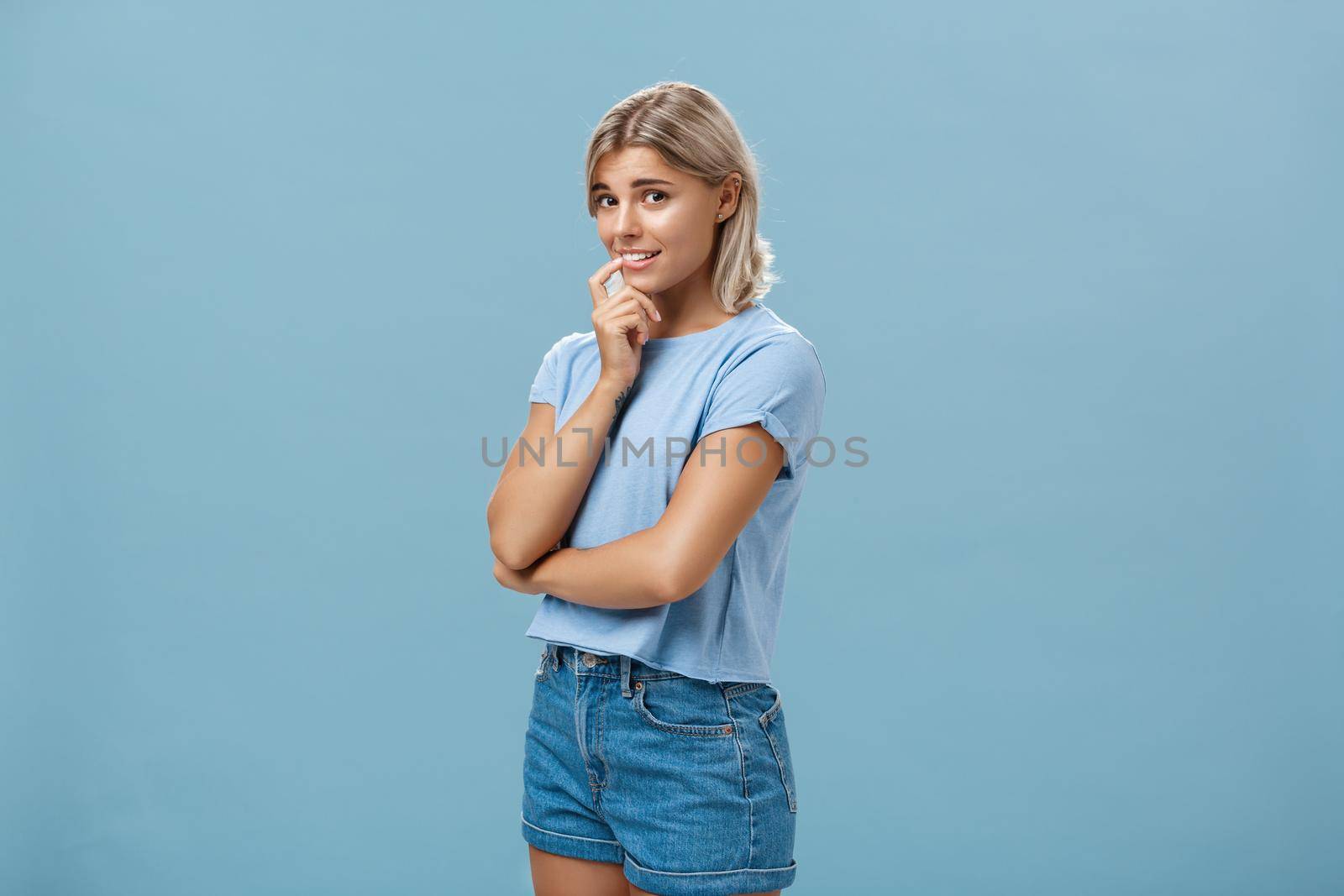 Studio shot of unconfident silly and insecure cute feminine girl with blond hair standing half-turned over blue background holding finger on lip while being shy ask question smiling hesitating by Benzoix