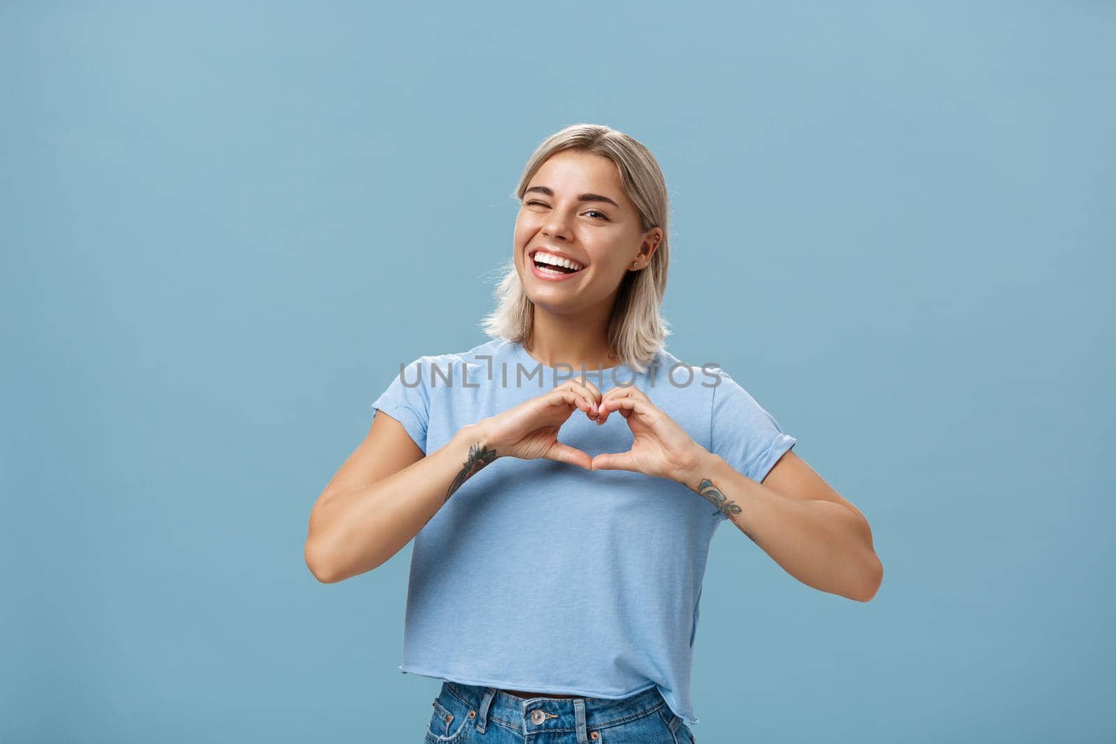 Love relationship and emotions concept. Joyful charming feminine girl with fair hair in trendy t-shirt winking happily smiling broadly and making heart gesture over breast expressing affection.