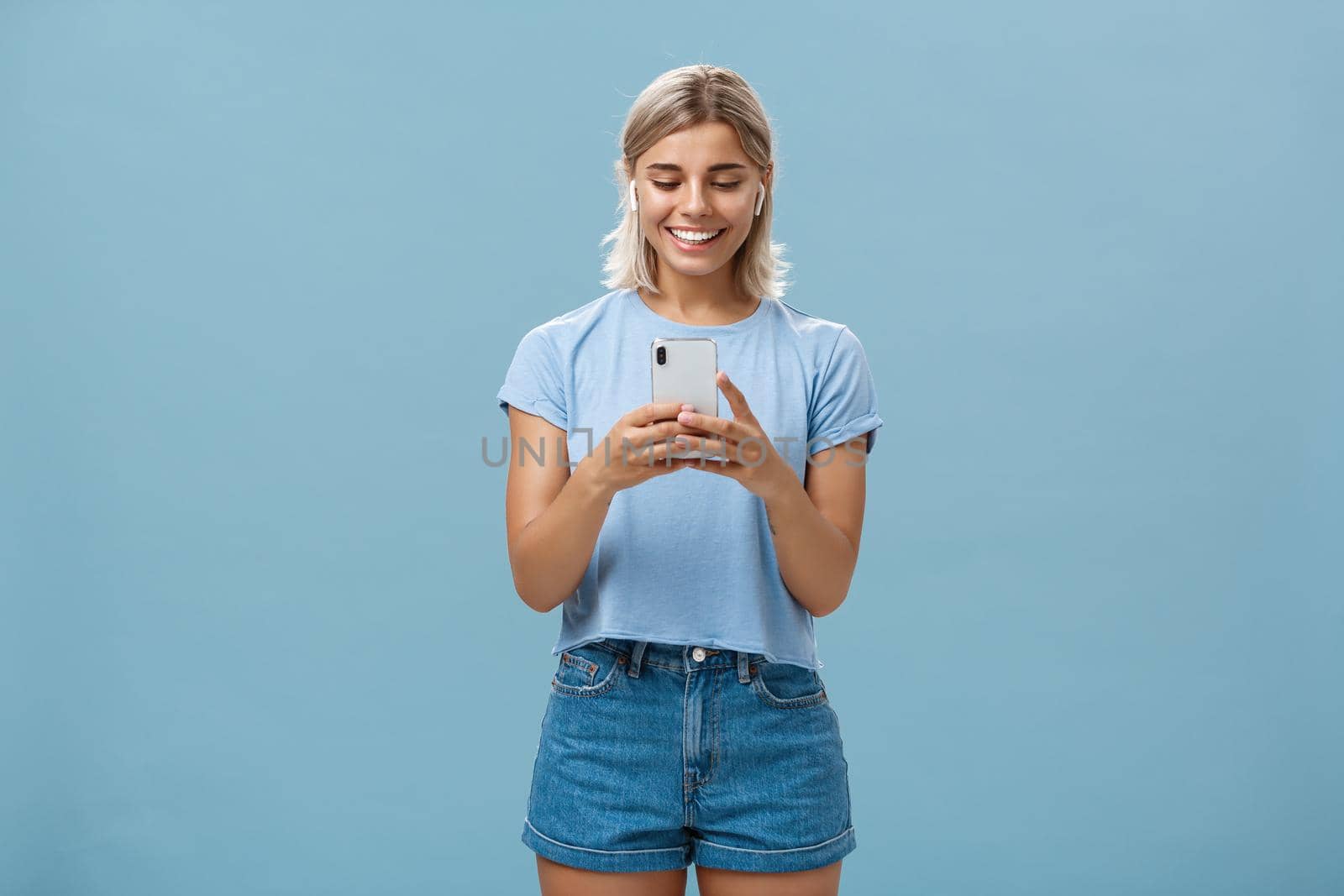 Girl having funny convesation, speaking with friend via video messages in wireless earphones holding smartphone in both hands smiling happily at device screen being entertained and amused. Emotions and technology concept