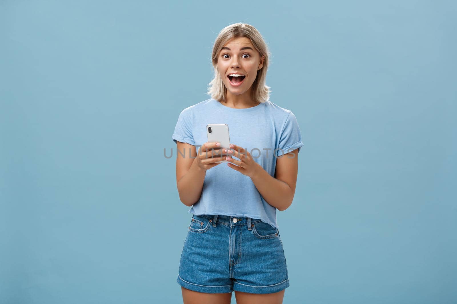 So happy receive premium for free reacting with enthusiasm and excitement on amazing message smiling broadly with delight and surprise holding smartphone posing over blue background. Copy space