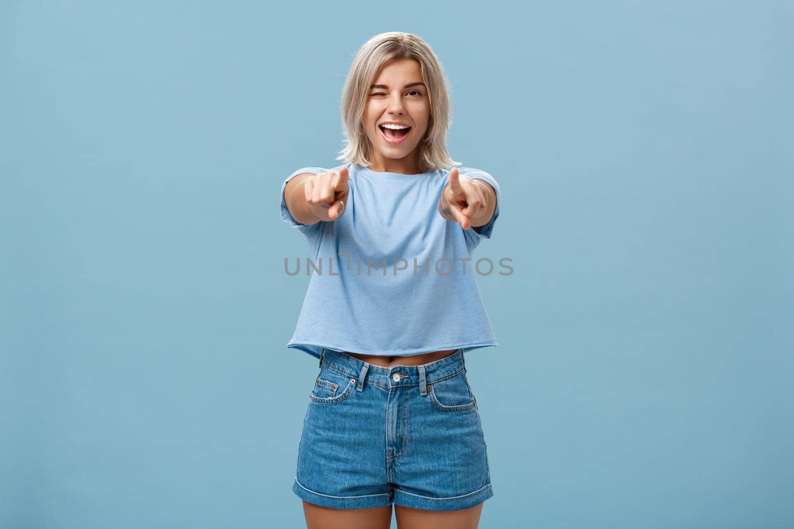 Happy enthusiastic stylish blonde female with tattoos pointing at camera as if picking or making hint smiling joyfully and winking from amazement and happiness posing over blue background. Copy space