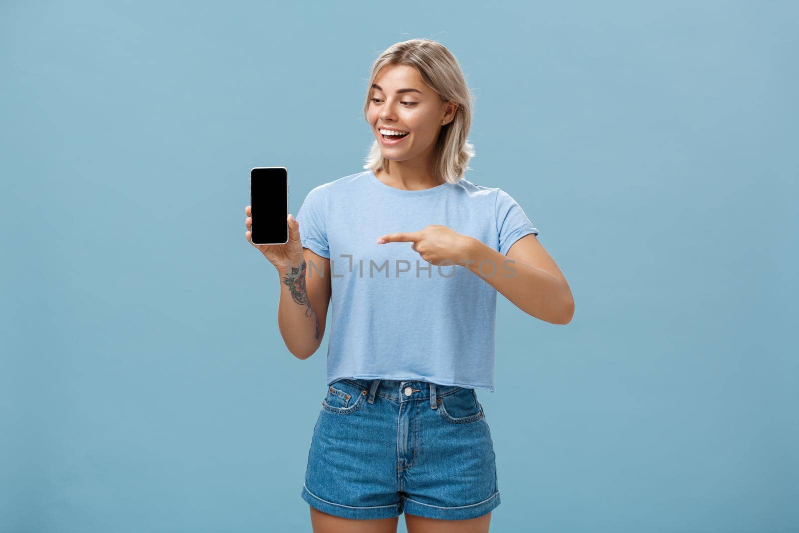 Portrait of delighted good-looking european female with blond haircut showing smartphone screen with amazement and delight pointing at device while promoting brand new gadget over blue background by Benzoix