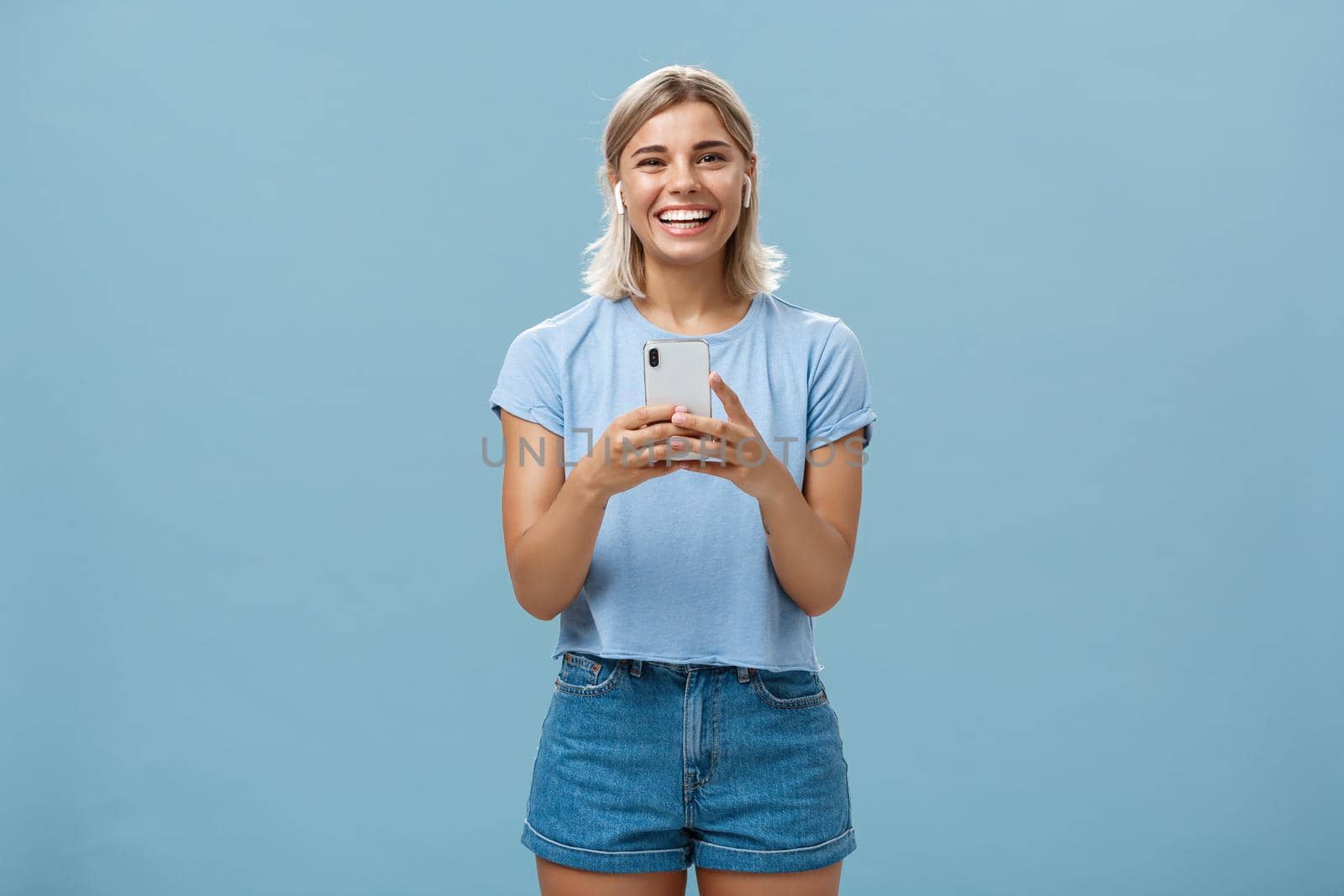 Satisfied happy and carefree young attractive blonde woman in casual outfit smiling broadly from happiness and delight listening music in wireless earphones holding smartphone over blue wall. Technology and lifestyle concept