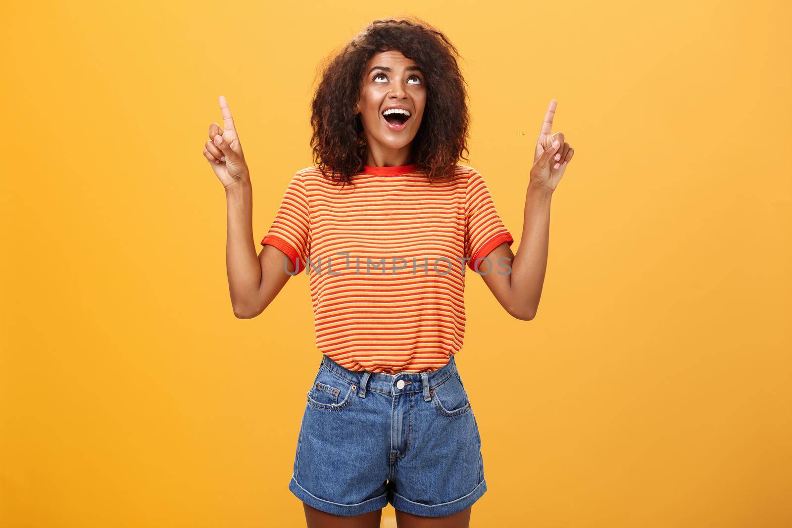 Fascinated impressed and amused good-looking charming African-American woman with afro hairstyle in trendy t-shirt and denim shorts pointing and looking up with interested look over orange wall. Lifestyle.