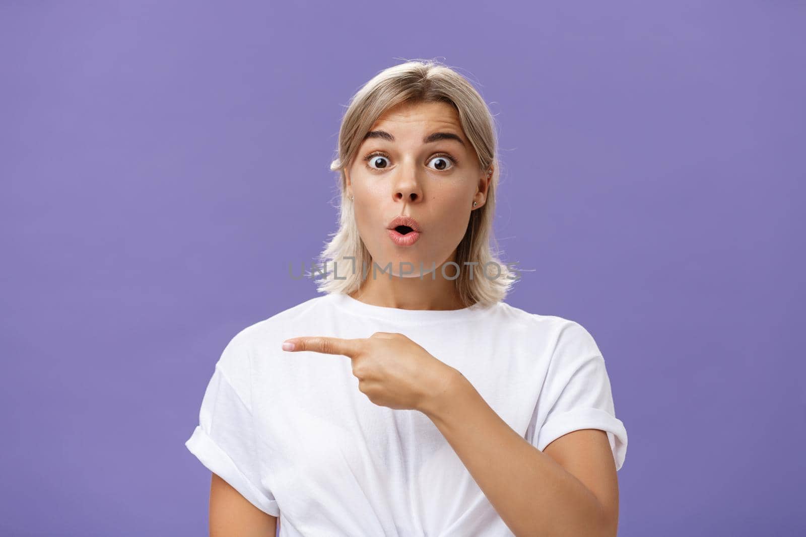 Waist-up shot of overwhelmed excited and surprised attractive urban female in white t-shirt folding lips gasping from amazement being questioned asking question and pointing left over purple wall. Copy space