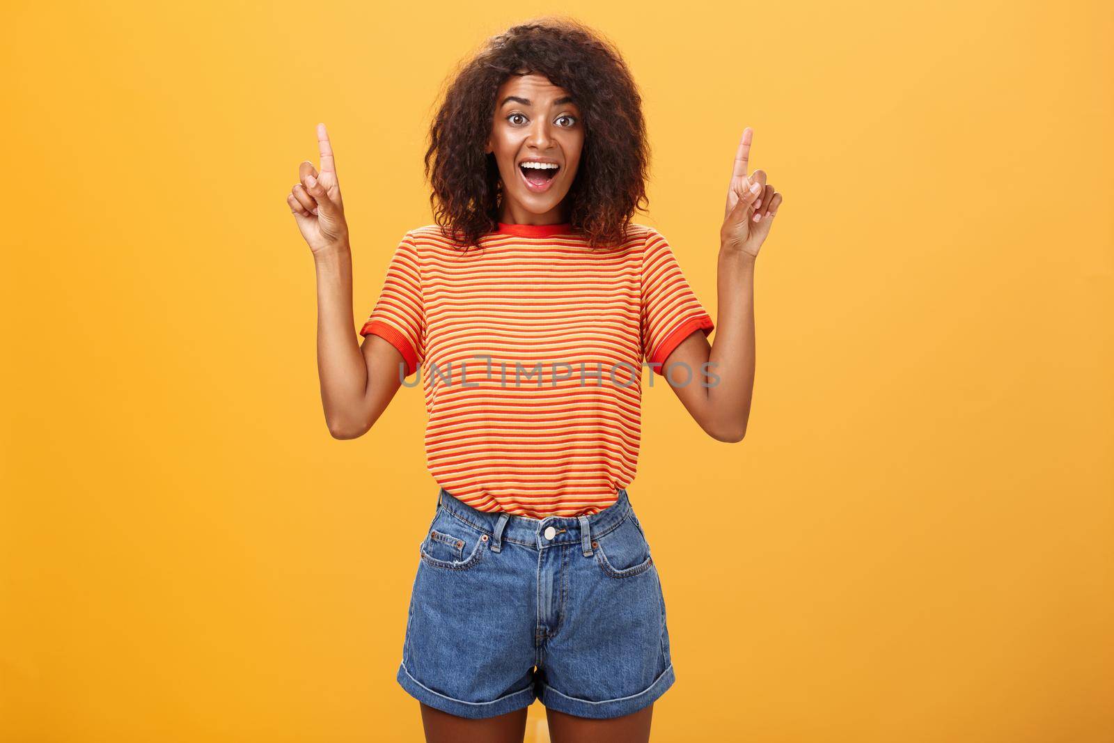 Charmed and impressed african american stylish woman seeing incredible and awesome item on sale raising hands and pointing up with broad grin being excited of new purchase over orange wall. Emotions and advertisement concept