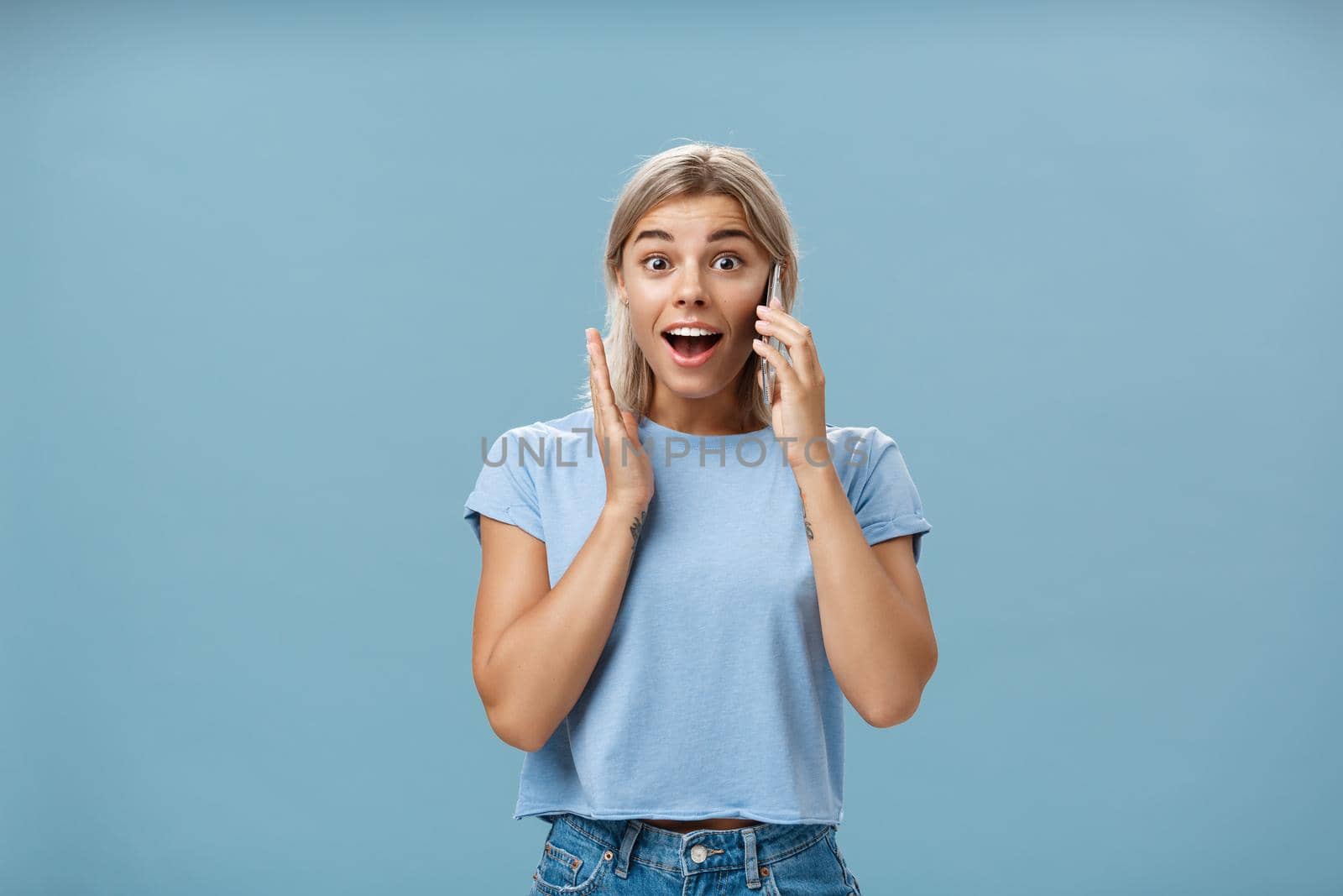 Pleased to hear best friend calling from abroad. Portrait of surprised pleased charming blonde female in trendy t-shirt opening mouth from amazement holding smartphone near ear and hand on face. Technology concept