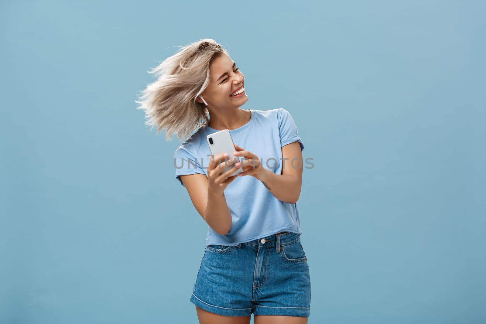 Great vibes carrying me away. Carefree joyful attractive young blonde female in stylish outfit waving head and smiling broadly listening music in wireless earbuds and holding smartphone over blue wall. Emotions and technology concept