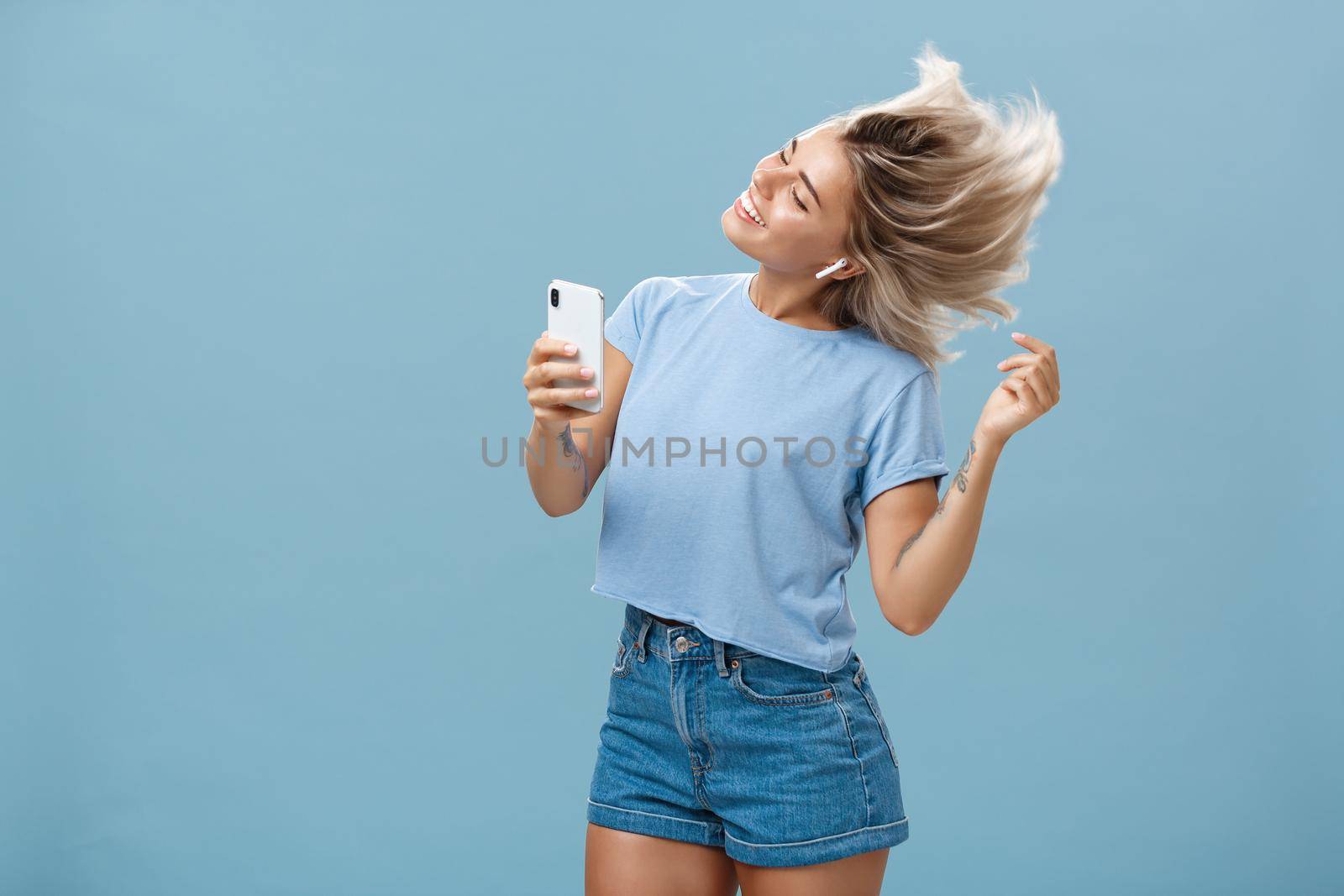 Lifestyle. Girl enjoying cool bits in brand new wireless earphones advertising earbuds in own blog recodring video via smartphone dancing from joy and delight smiling listening music over blue background.