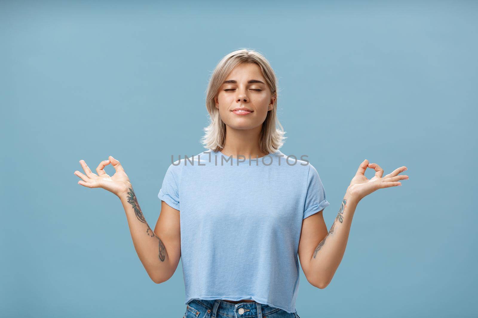 Listening to inner self during meditation. Calm happy and relaxed attractive tanned blonde female with tattoos on arms standing in lotus pose over blue wall with zen gesture closed eyes doing yoga by Benzoix