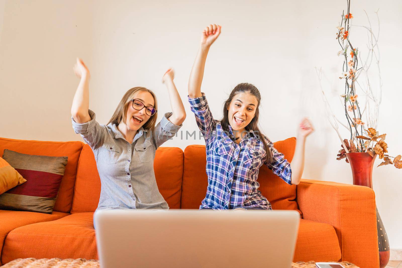Two very happy girl cheering with hands up looking at laptop screen sitting at home sofa. Young winner women having success in their work. Change your life and make preferred job to live better by robbyfontanesi