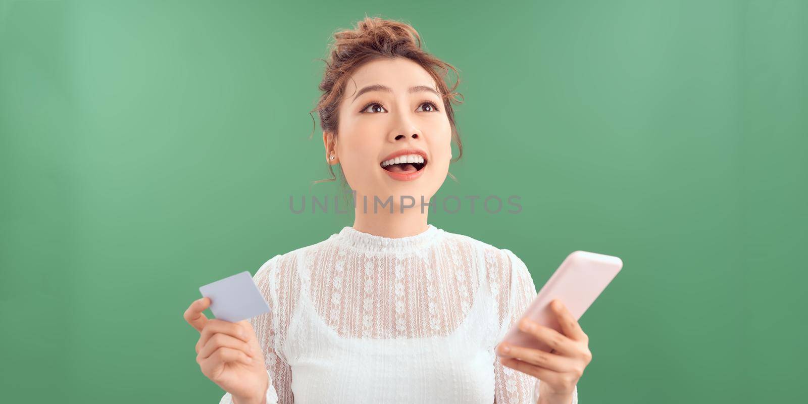 Photo of pleased young woman posing isolated over green wall background using mobile phone holding debit card.