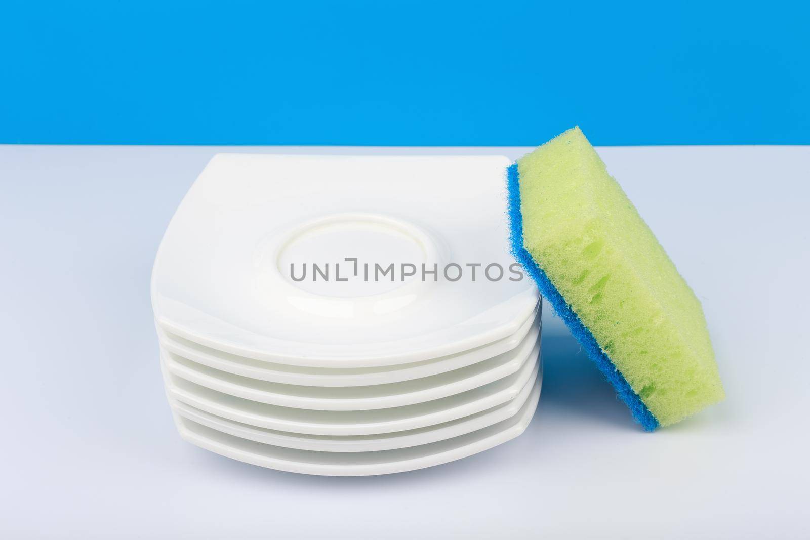 Dishwashing concept, high angle view of cleaning sponge and pile of clean saucers on white table with blue background. Stack of clean plates and dishwashing sponge close up