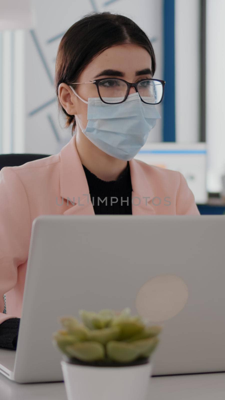 Close up of freelancer talking with coworker analaysing business statistics while wearing protective face mask. Team sitting in new normal office mantain social distancing during coronavirus pandemic