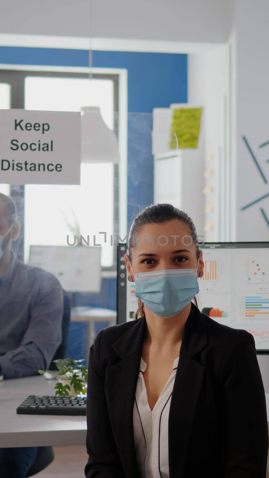Portrait of woman manager wearing face mask to prevent infection with coronavirus sitting on chair at desk table in business ofice. Colleagues maintain social distancing using separeted plastic board