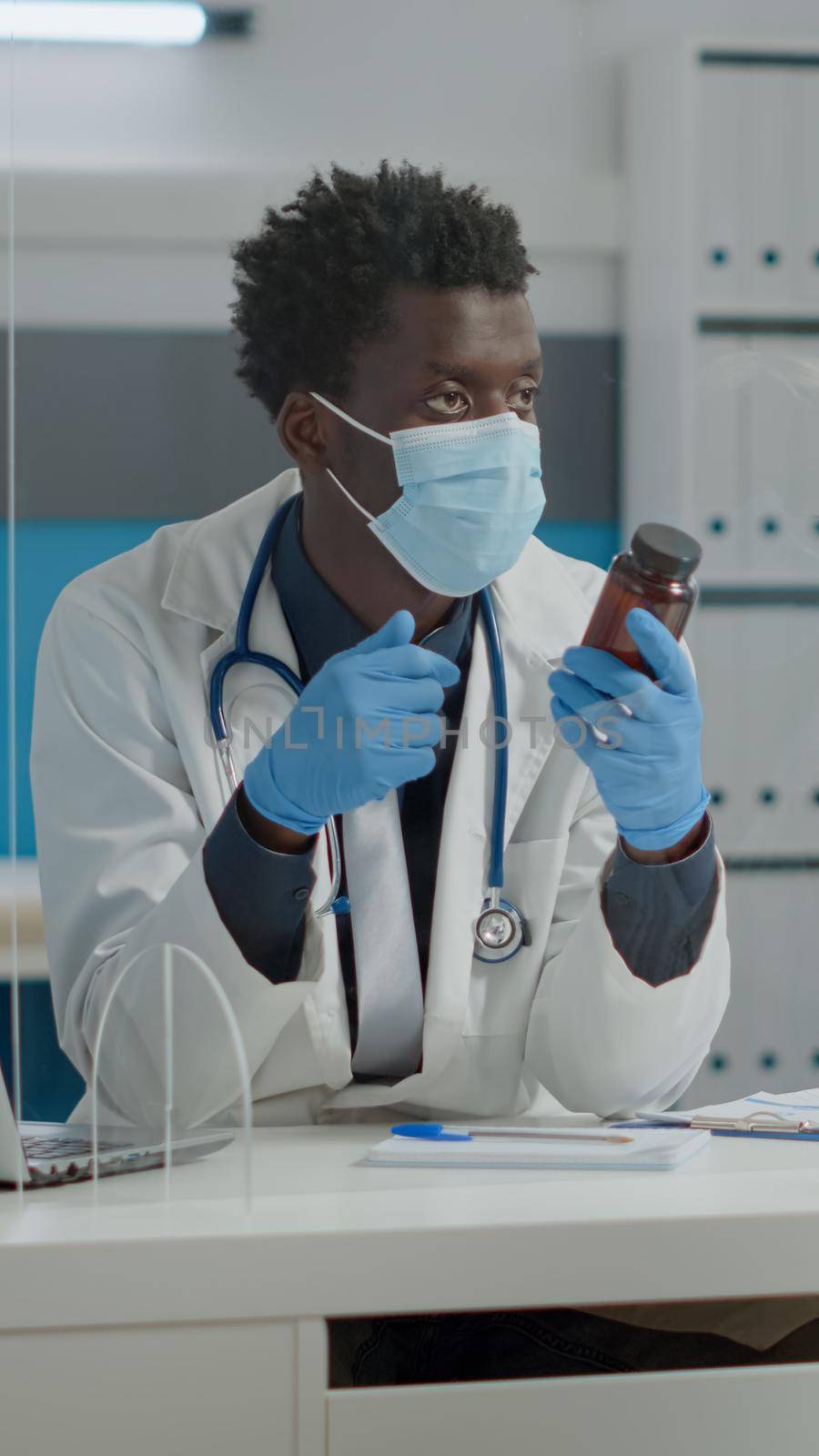 Young medic showing bottle of pills to elder patient for healing treatment in medical cabinet at healthcare clinic. Senior woman with disease receiving flask of medicine behind plexiglass wall