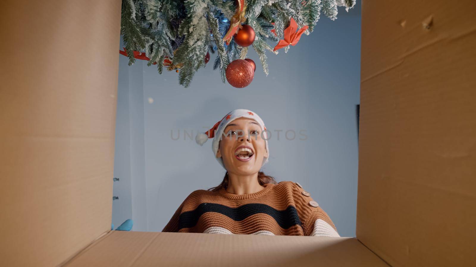 Young woman opening christmas present under tree being happy about surprise from friends and family. Caucasian person unpacking gift box while preparing for celebration festivity
