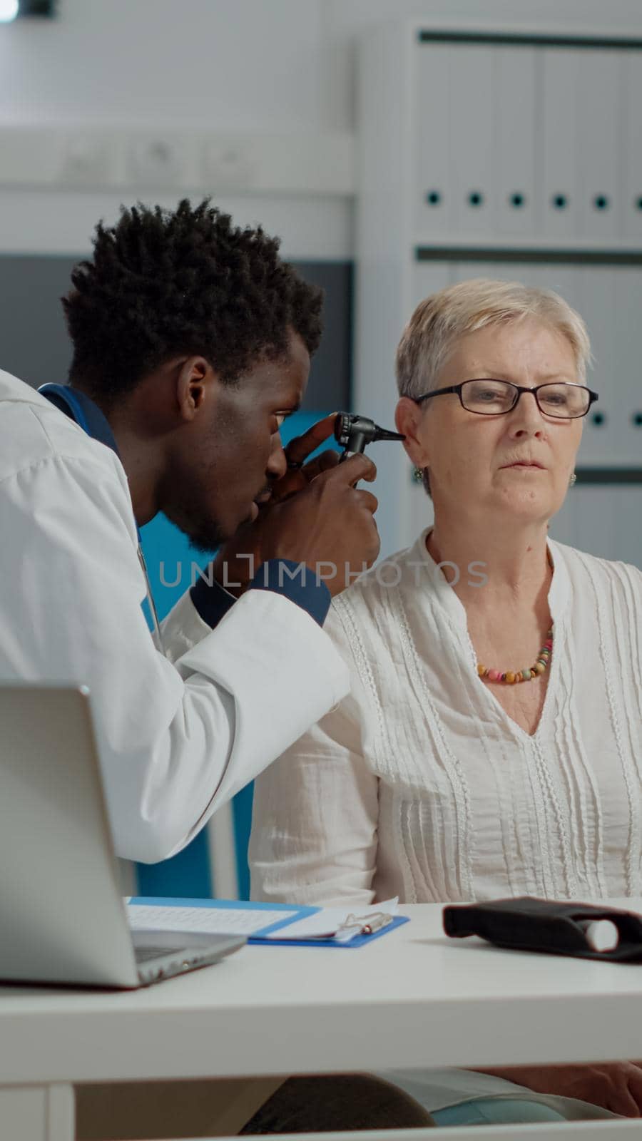 Young doctor using otoscope for ear checkup in cabinet with senior person at medical facility. Otologist doing examination with professional tool sitting in modern healthcare office