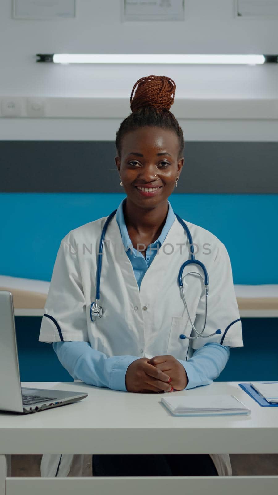 Portrait of african american woman with doctor profession wearing white coat and stethoscope at medical facility. Black medic smiling looking at camera in cabinet with nurse in background