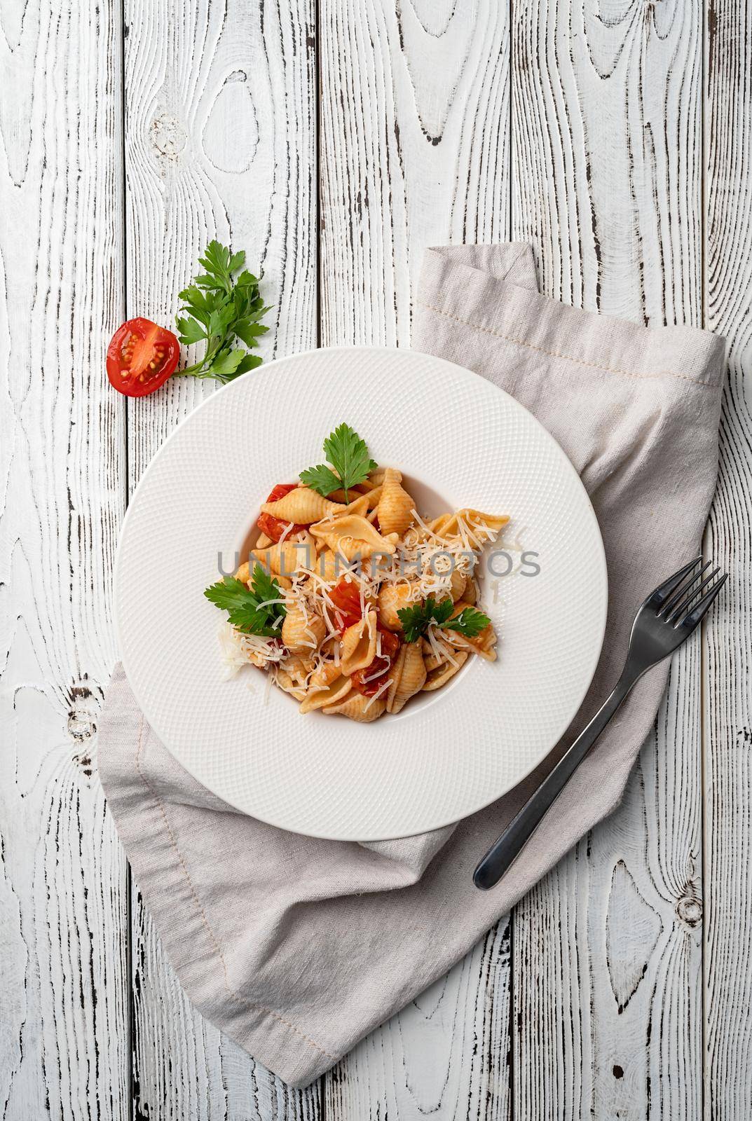 conchiglioni pasta with cherry tomatoes, cheese and parsley in white plate on wooden background, top view copy space