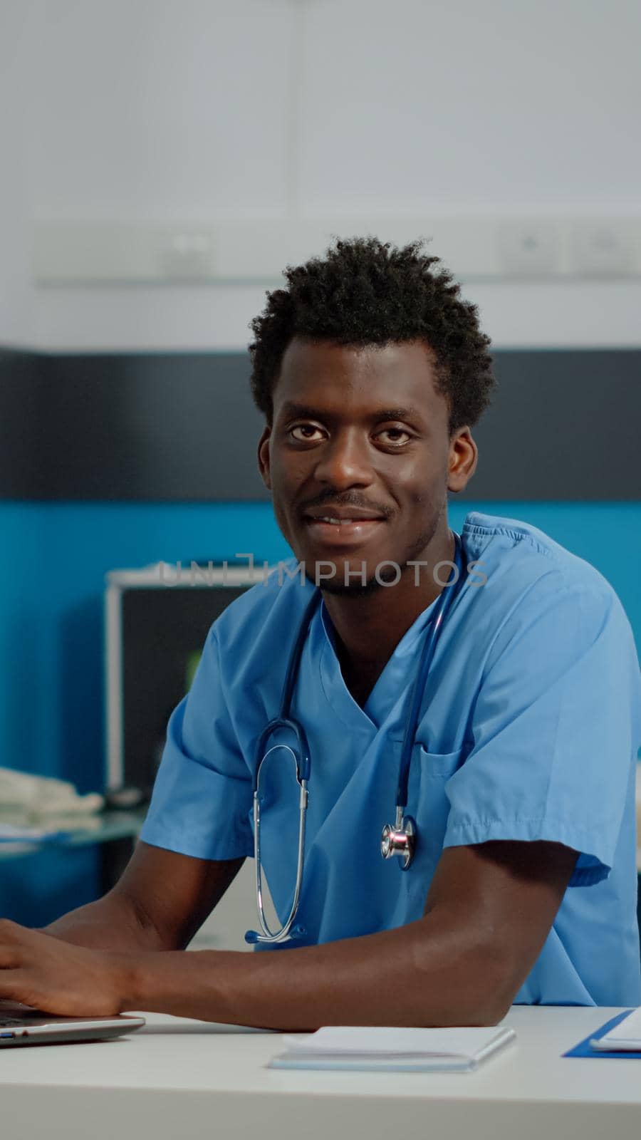 Portrait of male nurse with uniform and stethoscope using laptop on desk sitting in medical cabinet at facility. Healthcare assistant looking at camera, having modern technology
