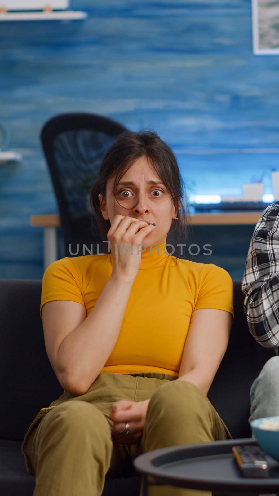 POV of shocked interracial couple watching scary movie on television at home. Multi ethnic husband and wife being afraid and covering eyes while looking at camera in living room
