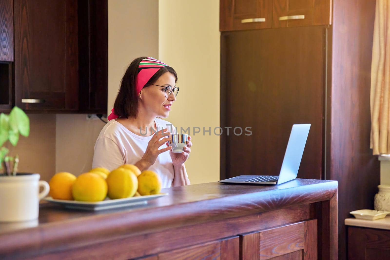 Woman in the kitchen with a cup of coffee looking into the laptop screen. Middle aged woman using video call for meeting and morning coffee. Technology, lifestyle, home, mature people concept