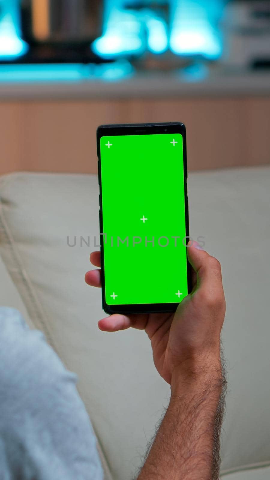 Relaxed man looking at smartphone with mock up green screen chroma key display while relaxing on sofa. Caucasian male holding in horizontal mode phone with isolated display late at night in kitchen
