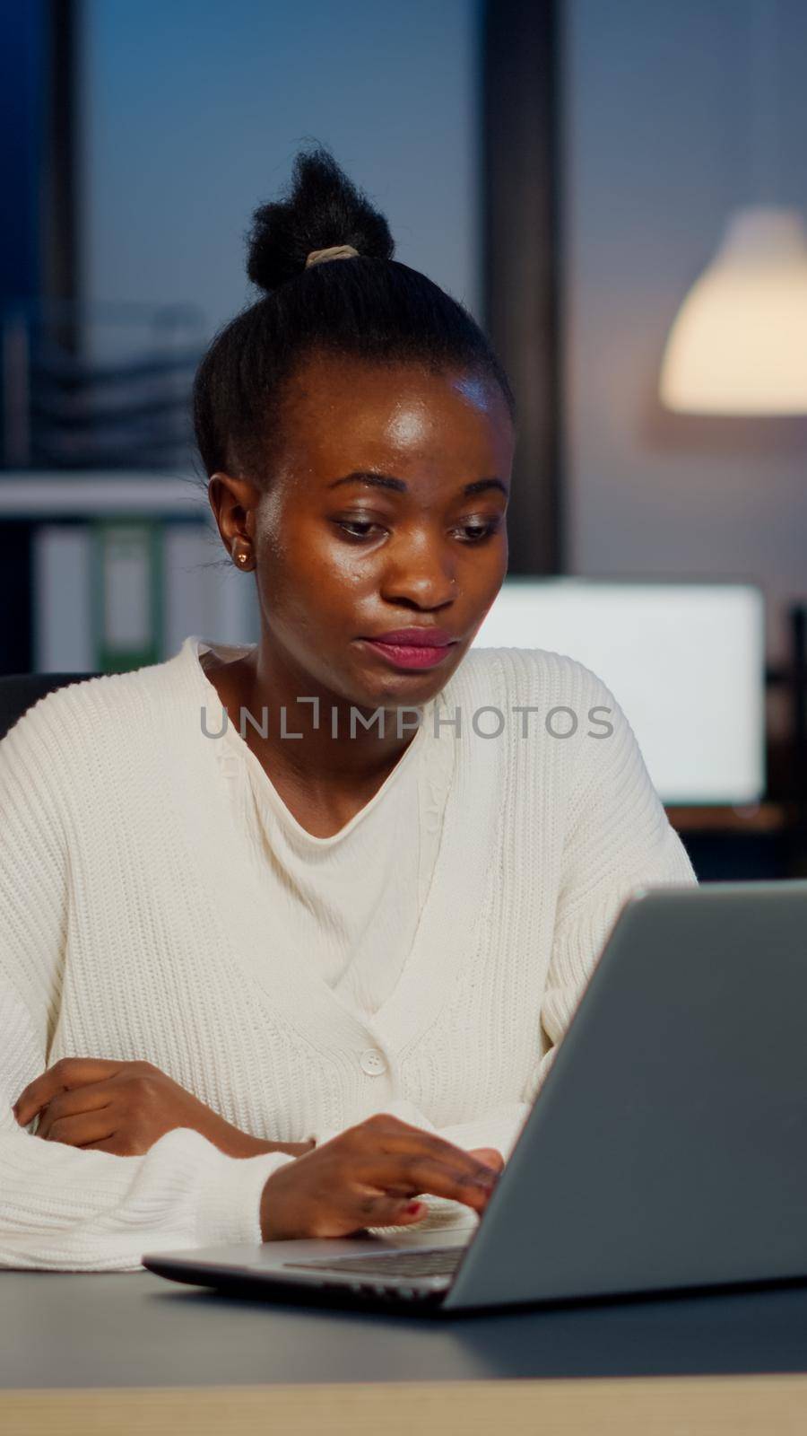 Stressed african manager woman working with financial graphs checking graphs, typing on laptop, reading raports late at night in start-up office doing overtime to respect deadline of project.