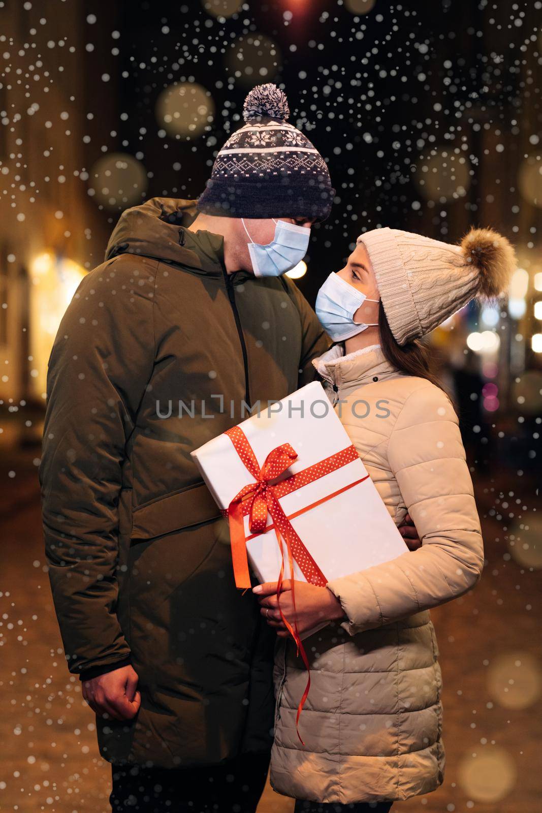 Xmas celebration together. Lovers travelling to Europe at the height. Couple in protective medical face masks celebrating xmas eve together at bright garland decoration. Christmas gifts exchange.