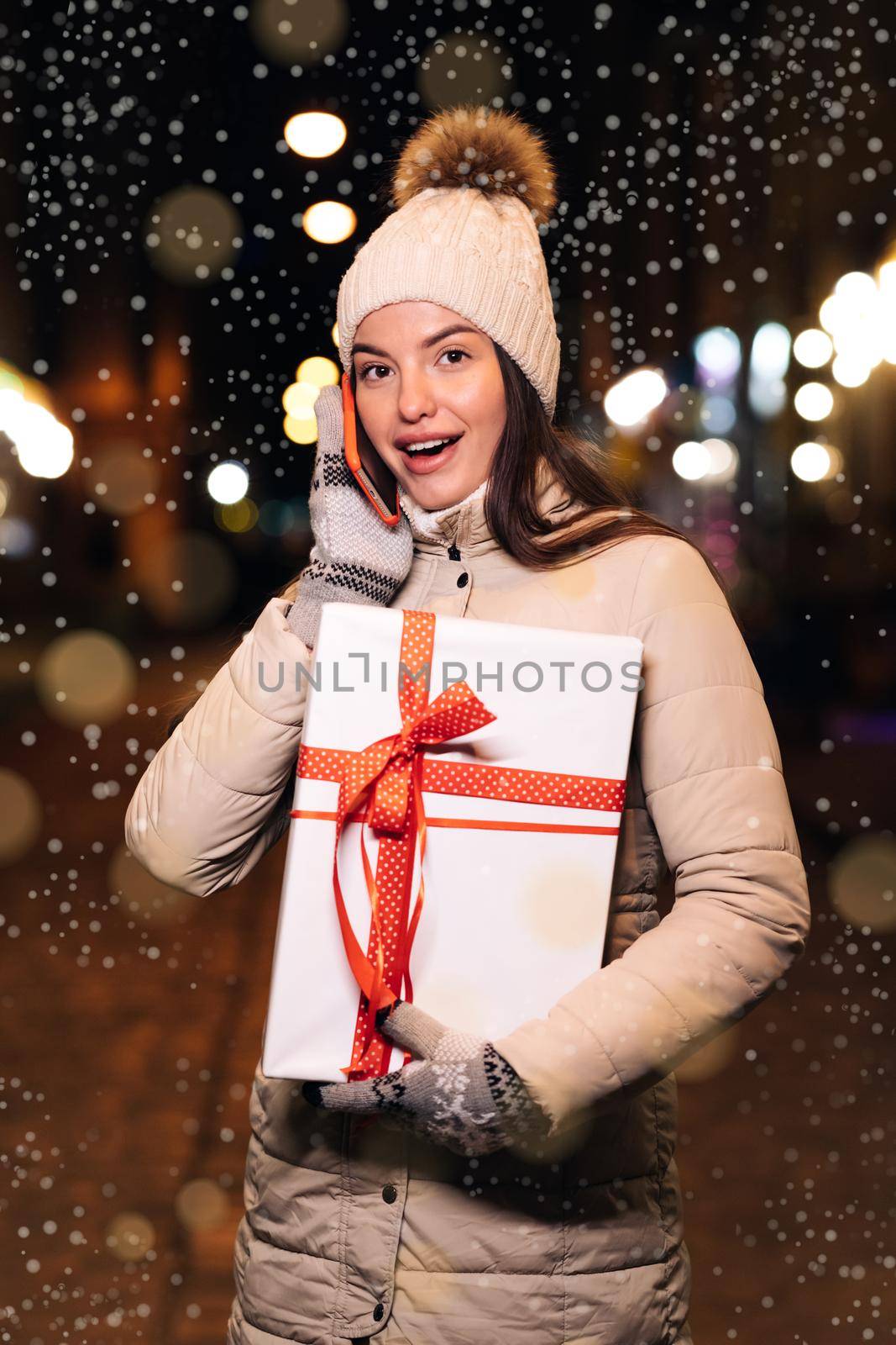 Vertical Shot of portrait beautiful female talking on smartphone while standing on street. Young woman with christmas present talking on cellphone. Winter concept. Vertical Screen Orientation