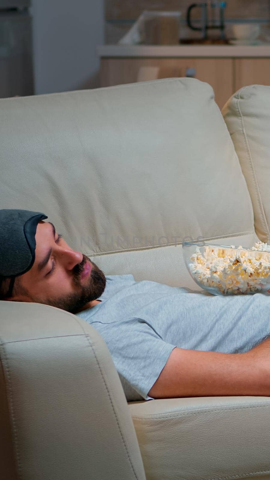 Man closing his eye and falling asleep on the couch in living room in front of the TV, while the television entertainment is ON. Relaxation cinema after a hard week of work