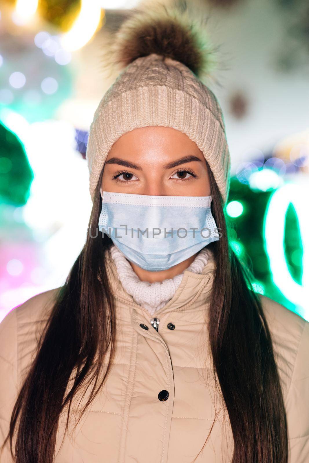 Vertical Shot of Close up portrait of Caucasian young female in medical face mask standing in decorated xmas city. Crowd people sick. Epidemic coronavirus. Pandemic flu corona virus by uflypro