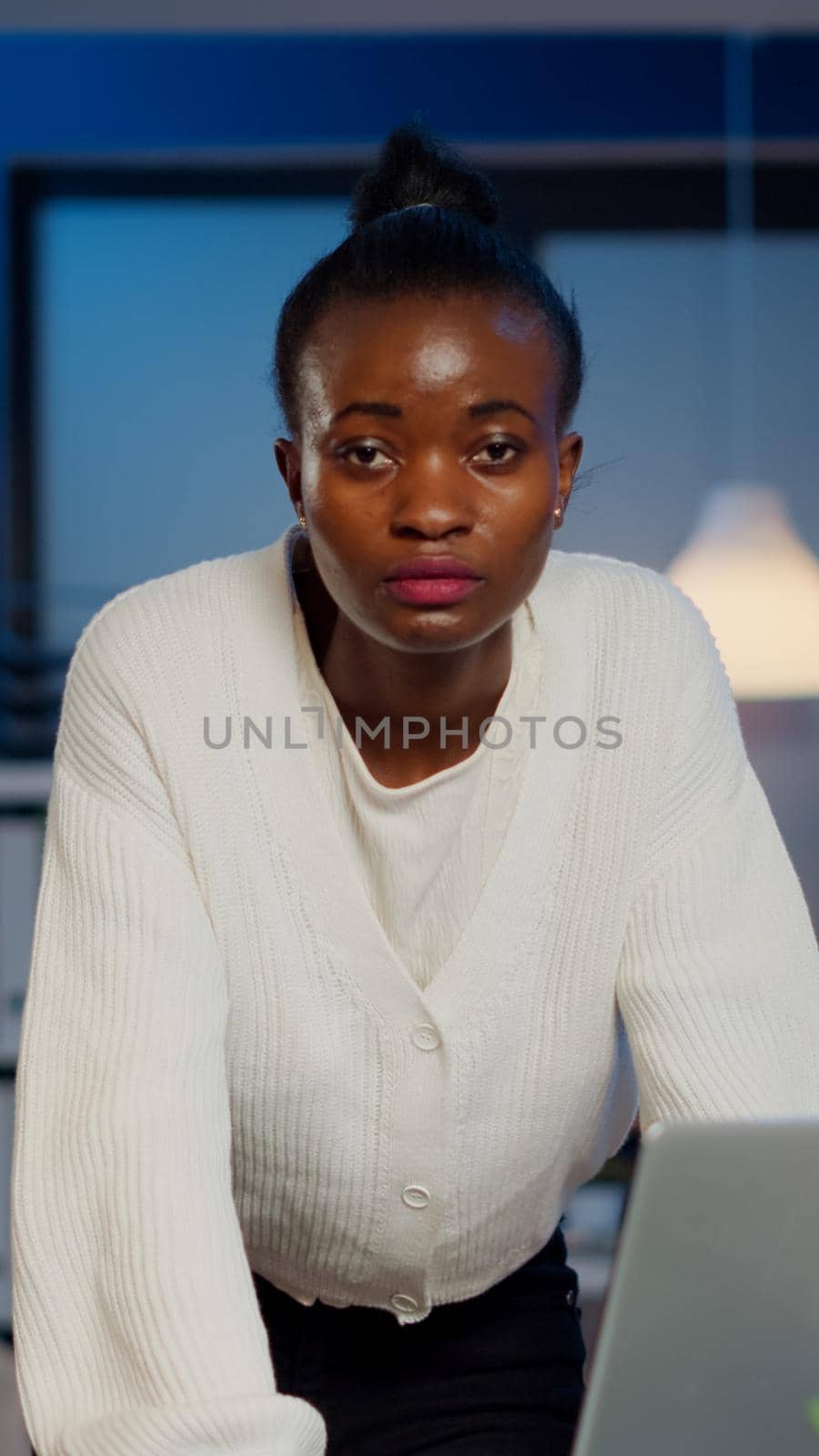 Focused tired business woman looking at camera after reading tasks on laptop standing near desk in start-up company late at night. Employee doing overtime for job respecting deadline