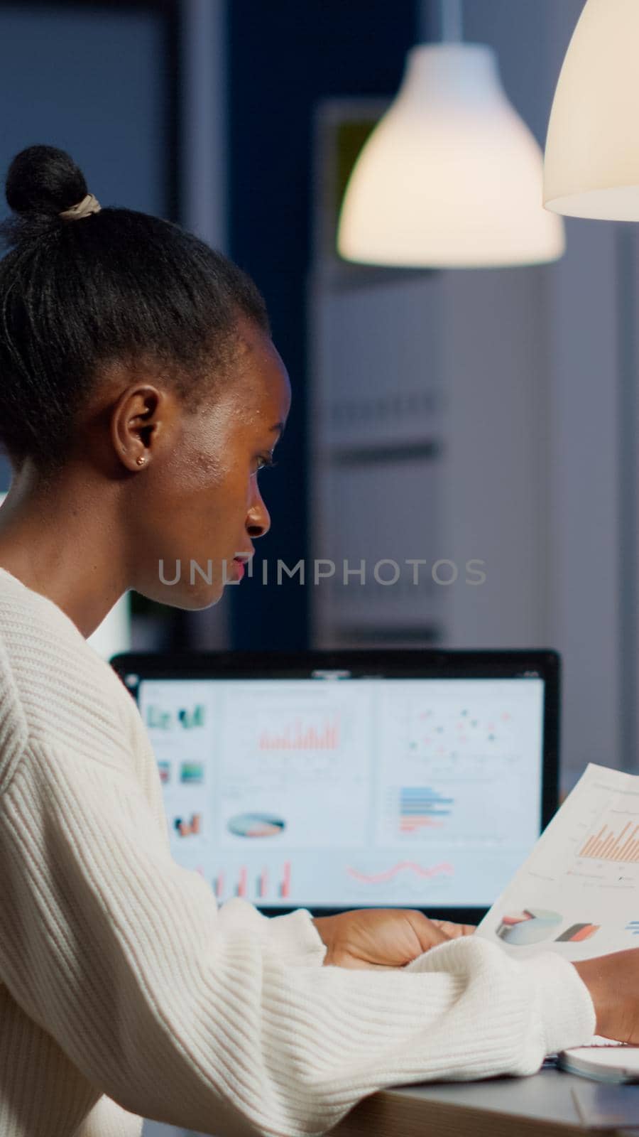 African business woman holding documents with graphics and financial statistics working overtime in front of laptop sitting in start-up business office. Focused employee respecting deadline of project