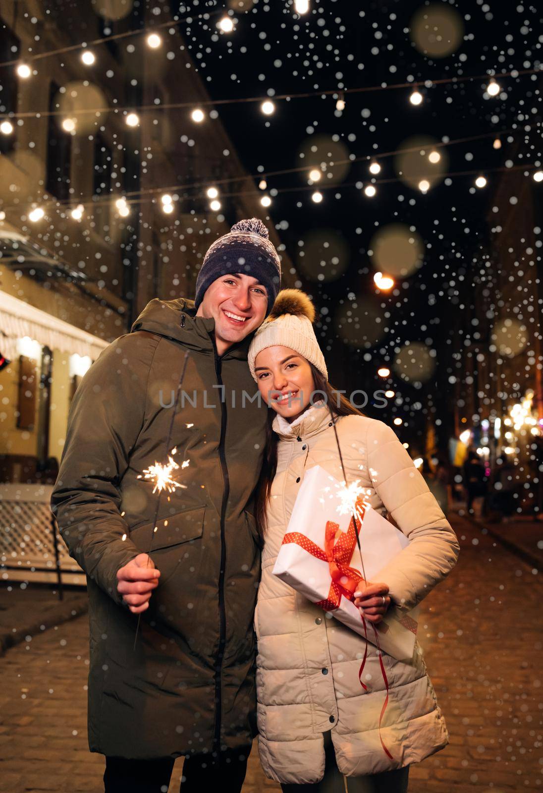 Romantic couple in love with gift box hugging and burning the sparklers. Lovers travelling to Europe at the height of Christmas holidays. Sweethearts in the centre of city.