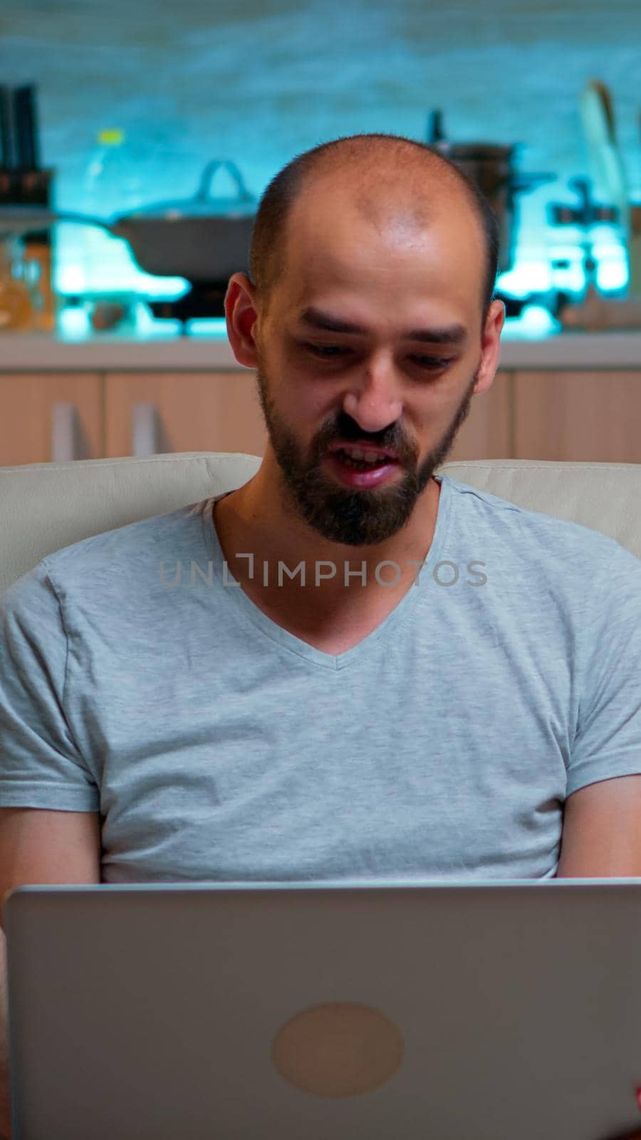 Caucasian male sitting on couch in pajamas working on laptop computer during videocall conference. Comfortable man relaxing on sofa chatting with business people late at night in kitchen