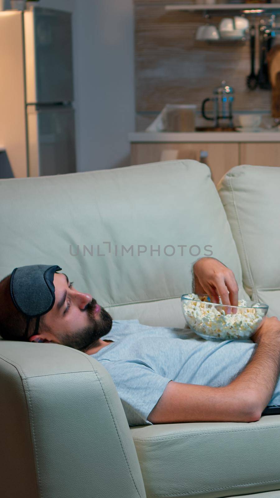 Man lying on sofa, falling asleep in fron of the TV with popcorn bowl in his hands. Tired alone man in living room after a hard day at work