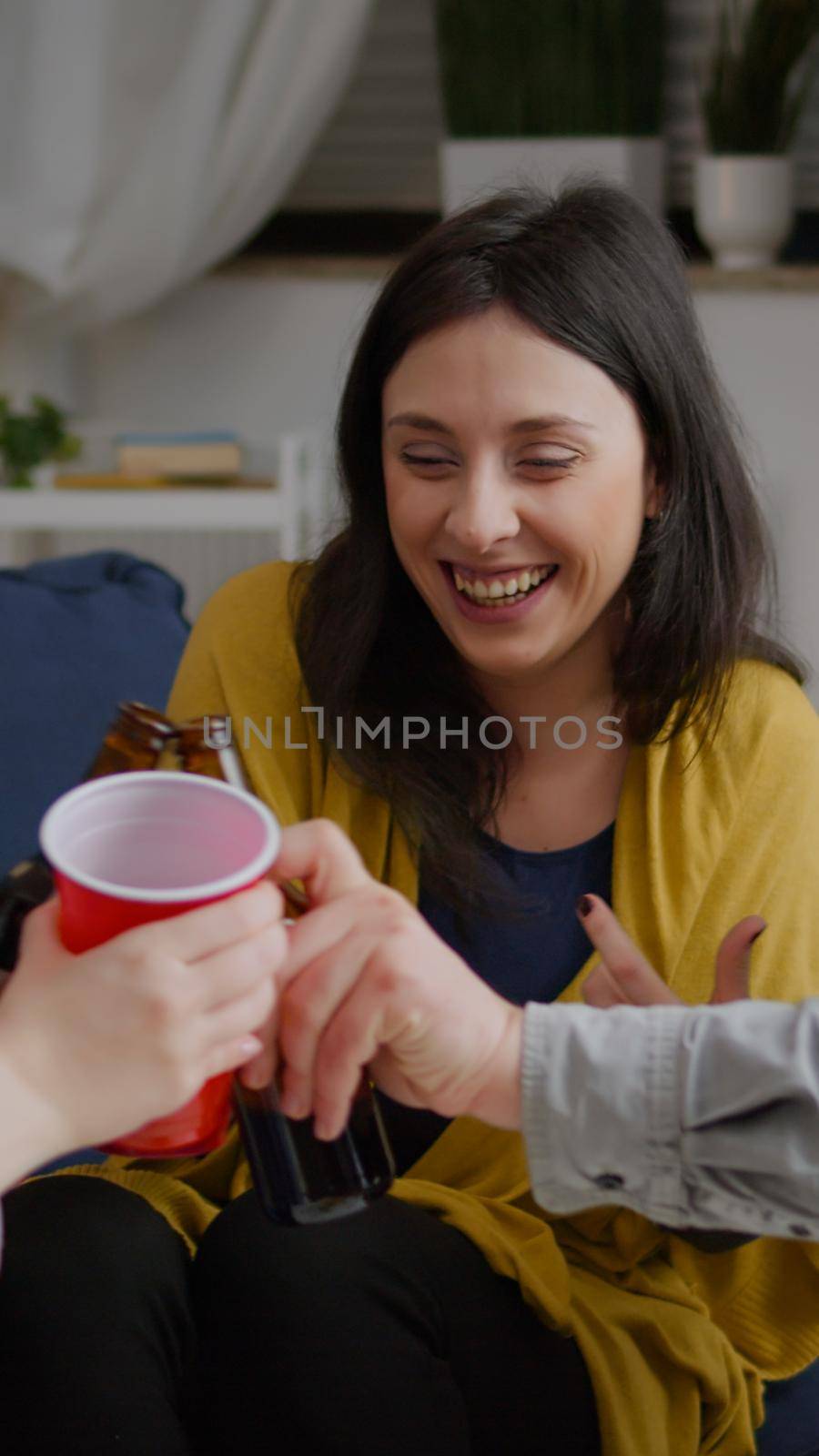 Multi-cultural friends socialising while hanging out late at night in living room during fun party. Group of mixed race people enjoying time spending together cheering beer bottles