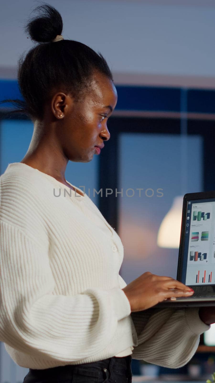Focused black manager woman checking business report standing in start up office late at night holding laptop. Busy african employee analysing financial statistics overworking writing, searching.